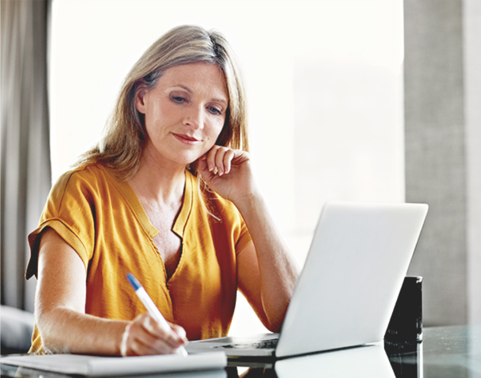 Femme assise à une table en verre, devant un ordinateur et prenant des notes